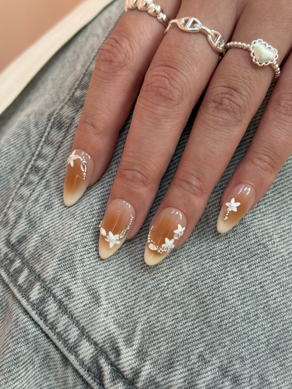 Hand with almond-shaped nails featuring floral designs, wearing rings on a denim background.